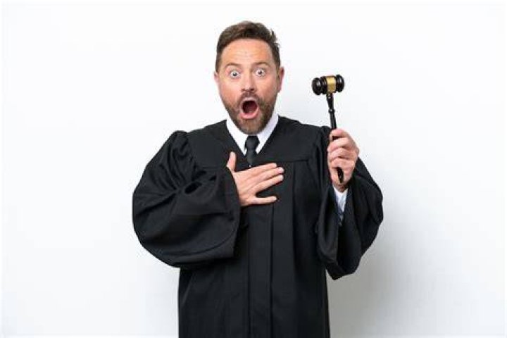 A man in judge's robes sits on a fancy leather chair in front of a bookshelf filled with law books.