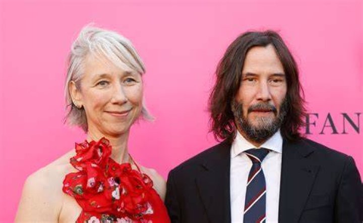Alexandra Grant and Keanu Reeves posing next to each other for a photo on the red carpet, standing in front of a black backdrop. 