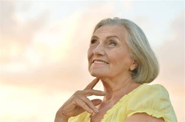 An older woman in a pink sweater vest combo leans on a countertop.